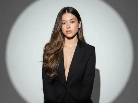 SashaCollins — Young woman in a chic black blazer and silver necklace posing against a minimalist grey studio background.
