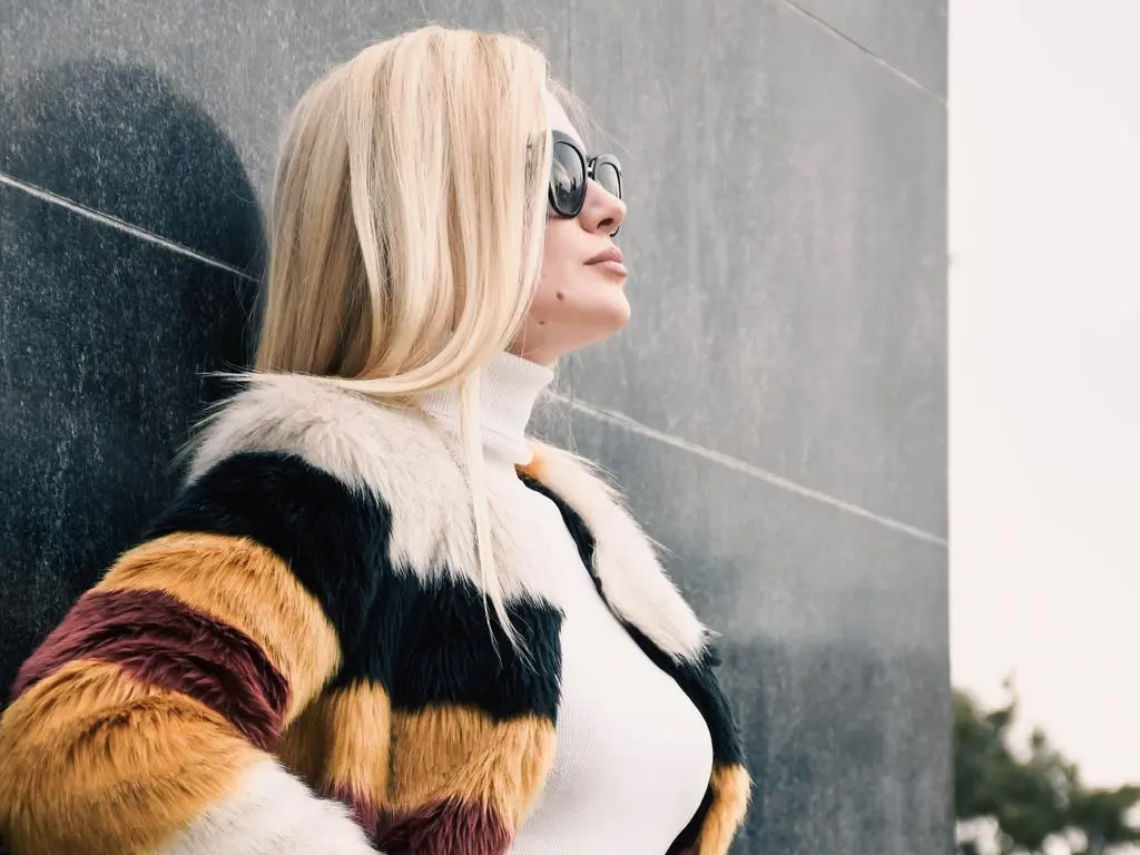 Blonde woman in a striped faux fur coat and white turtleneck posing against a dark stone wall.