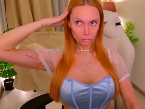 LoveCall — Redheaded woman in a blue satin corset top posing in a cozy, sunlit bedroom setting.