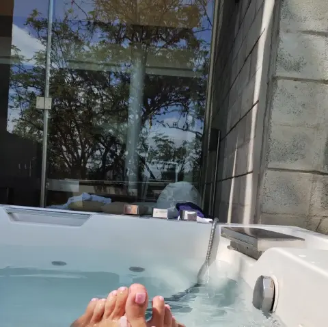 Hunny_Bunny relaxes in a white hot tub with her feet visible above the clear water against a glass window and stone wall.