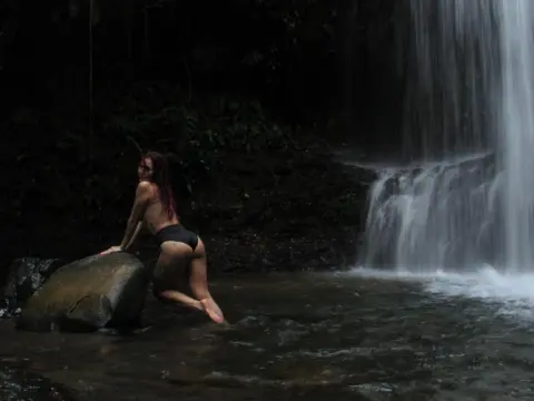 Hunny_Bunny poses naked beside a waterfall, kneeling on a large rock in a dark jungle stream.