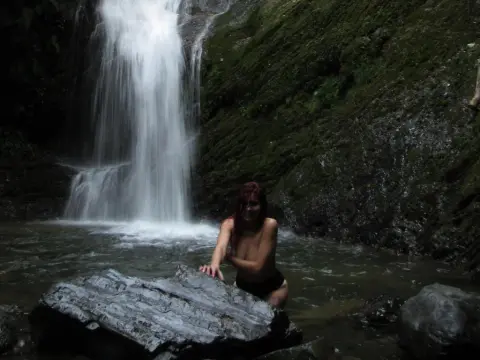 Hunny_Bunny sits naked in a dark pool of water in front of a waterfall, resting her hand on a large rock.