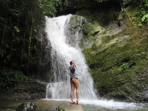 Hunny_Bunny stands in front of a waterfall, wearing a grey tank top and black undies with her hair pulled back.