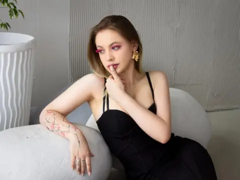 AuroraAmor poses in a black dress with her hand near her mouth, showing off her blonde hair and floral arm tattoo.