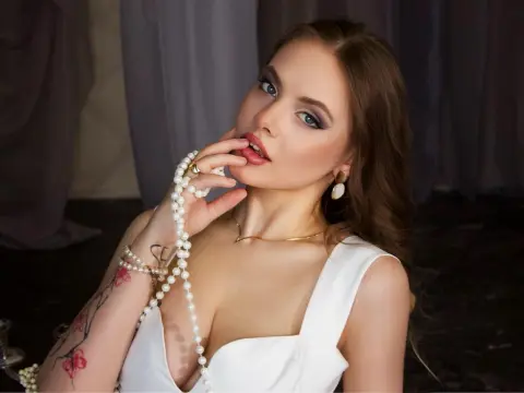 AuroraAmor poses with a pearl necklace against her chest, wearing a white low-cut top and gold jewelry.