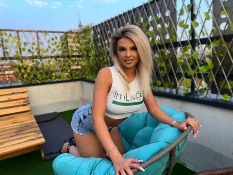 KimmyBliss — Blonde woman in white crop top and denim shorts sitting on a turquoise lounge chair on a sunny rooftop terrace.