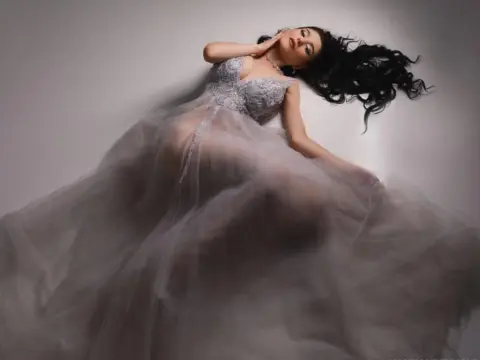 NorraJoice — NorraJoice lies on her back in a sheer grey lace gown with her long dark hair spread out.