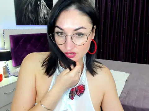 StefanaMurphy — Young woman with dark hair and glasses wearing a white halter top in a cozy bedroom setting.