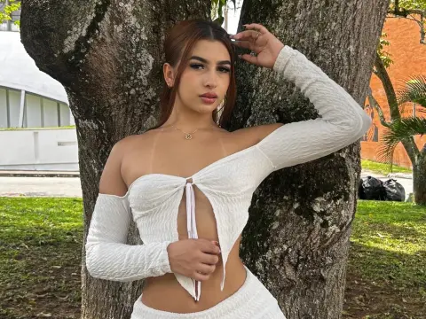 NicoleAdams_ — Young woman in a white off-the-shoulder crop top posing by a large tree in a sunny outdoor garden setting.