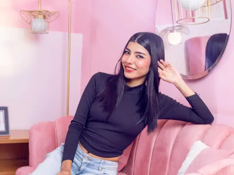 Smiling young woman in a black turtleneck sitting on a pink velvet sofa in a bright, pastel room.