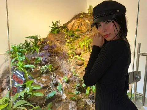 Young woman in a black ribbed sweater and cap posing against a natural rock wall with green plants.