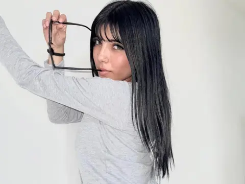 Young woman with long black hair and bangs wearing a grey long-sleeve shirt holding glasses against a white background.