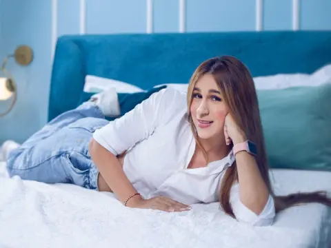 Smiling young woman in a white shirt and jeans relaxing on a blue bed in a bright, cozy bedroom.