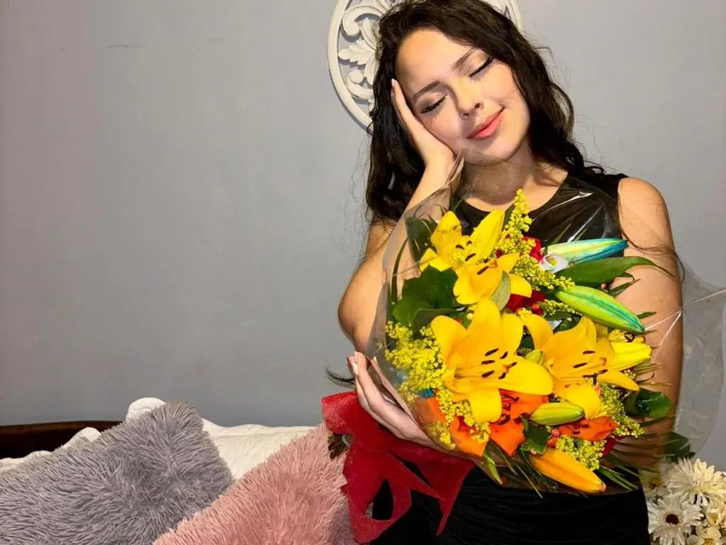 Alison_Tatcher — Smiling young woman in a black dress holding a bright yellow lily bouquet in a cozy bedroom setting.