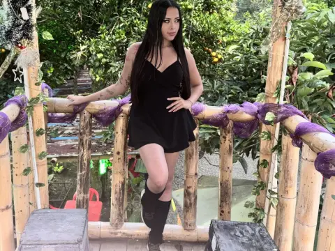 Alison_Tatcher — Young woman in a black summer dress standing on a bamboo balcony surrounded by lush green garden trees.