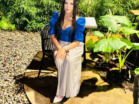 Alison_Tatcher — Young woman in a blue puff-sleeve top and beige trousers sitting at an outdoor garden cafe table.
