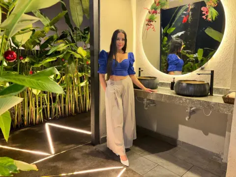 Alison_Tatcher — Woman in a blue puff-sleeve crop top and beige trousers standing in a tropical luxury bathroom at night.