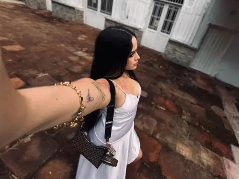 Alison_Tatcher — Tattooed woman in a white summer dress carrying a black handbag on a rustic cobblestone street.