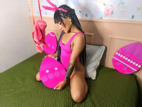 Young woman in a pink bodysuit sitting on a green bed with pink Easter eggs and a bunny plushie.
