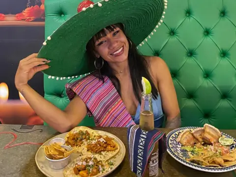 Smiling woman in a colorful striped top wearing a large green sombrero at a Mexican restaurant with tacos and beer.