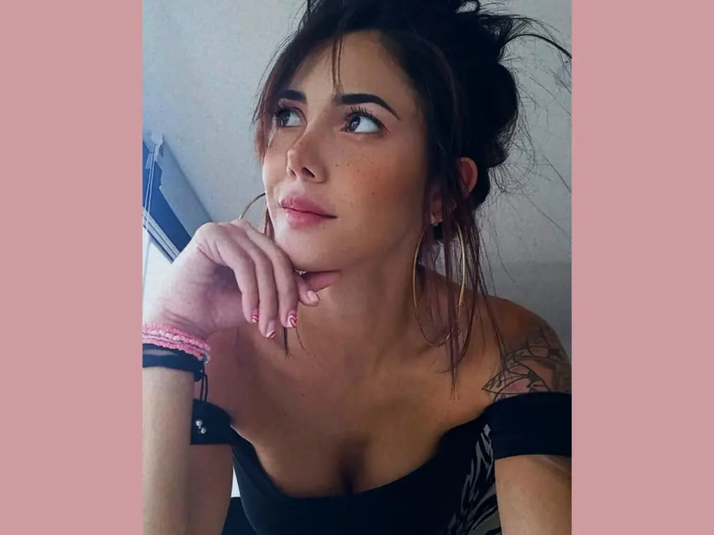 Mix_Kitty — Thoughtful young woman with messy bun and gold hoop earrings wearing a black off-the-shoulder top.