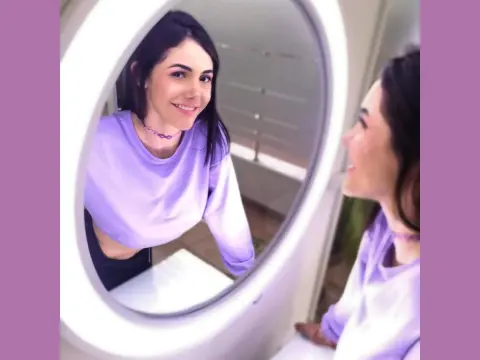 Mix_Kitty — Smiling young woman in a lavender long-sleeve crop top looking into a round mirror in a bright room.