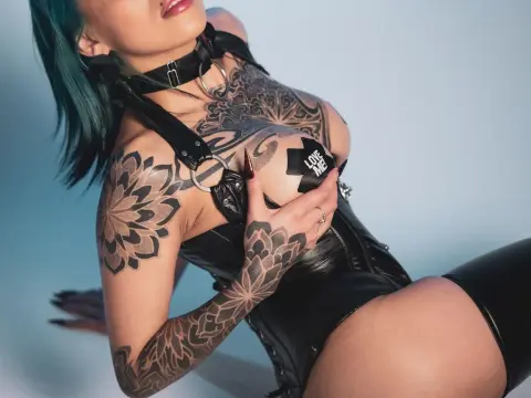AlissaPeach — Tattooed woman in black leather fetish outfit posing against a blue background with a bold, edgy mood.