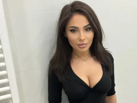 SarahSkye895 — Glamorous brunette woman in a black v-neck top posing against a white wall with a confident expression.