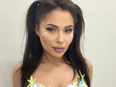 SarahSkye895 — Woman with colorful gem face makeup and pigtails wearing a pastel floral outfit in a bright setting.