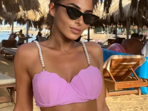 SarahSkye895 — Stylish woman in a lilac bikini top and black sunglasses relaxing at a sunny tropical beach resort.
