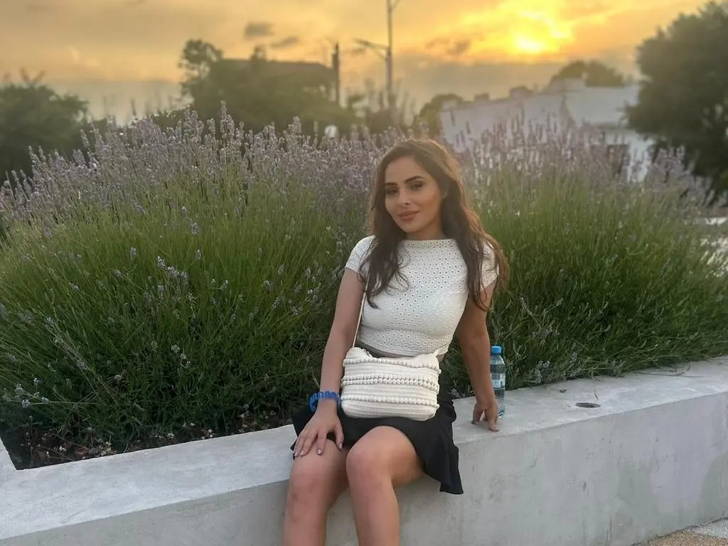 SarahSkye895 — Woman in white knit top and black skirt sitting by lavender bushes during a golden sunset.
