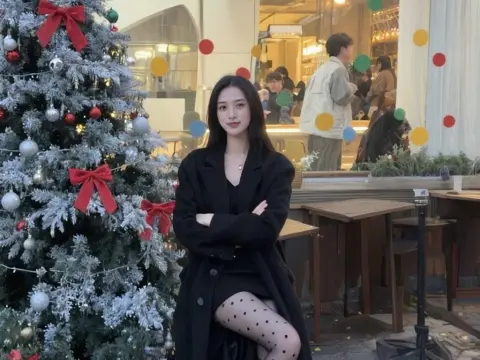 Young woman in a black coat posing by a decorated Christmas tree in a cozy cafe setting.