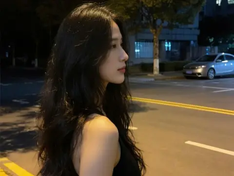 Young woman with long wavy hair in a black tank top standing on a city street at night.