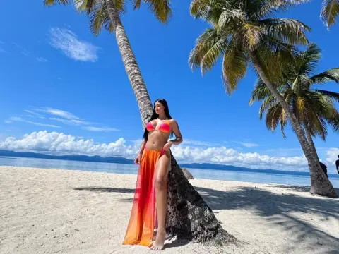 Woman in orange bikini and long sheer skirt posing by palm trees on a sunny tropical beach.