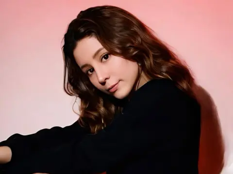 SweetTulip — Young woman with wavy brown hair wearing a black top against a soft pink studio background.