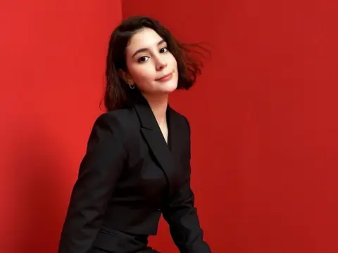 SweetTulip — Smiling young woman in a professional black blazer posing against a vibrant red studio background.