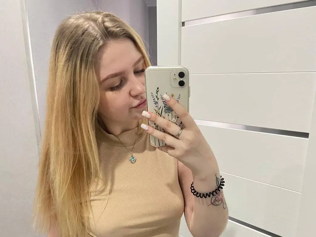 AnitaVine poses for a mirror selfie, wearing a tan ribbed tank top and a small heart pendant necklace, with her long blonde hair down.