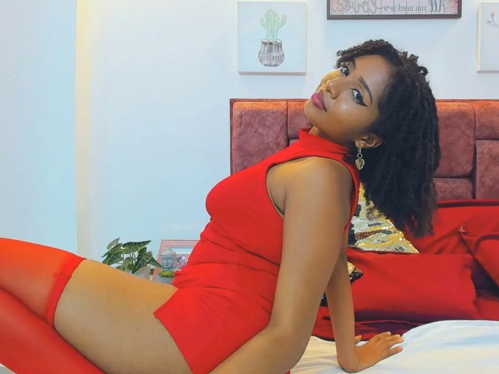 Megan_Wlliams — Beautiful woman in a red sleeveless dress lounging on a bed in a bright, cozy bedroom setting.