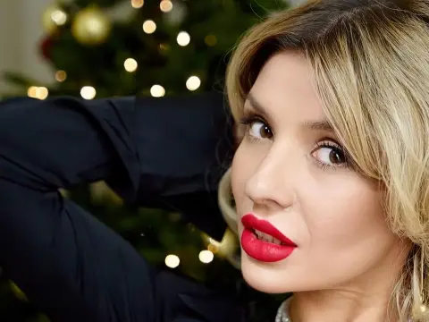MonaMia — Blonde woman in a black outfit with red lipstick posing against a festive, glowing Christmas tree background.