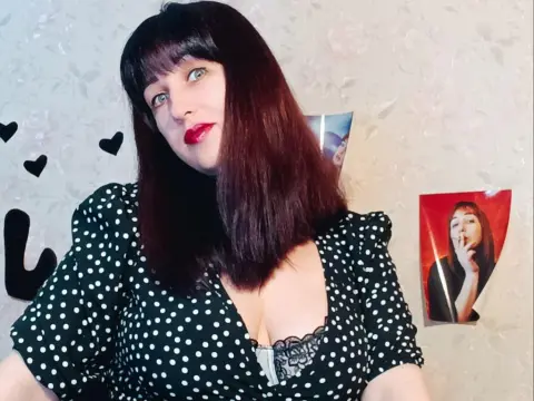 Woman with dark hair and red lipstick wearing a black and white polka dot dress in a cozy room.