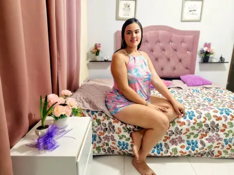 Smiling woman in a colorful floral halter dress sitting on a bed in a bright, cozy bedroom.