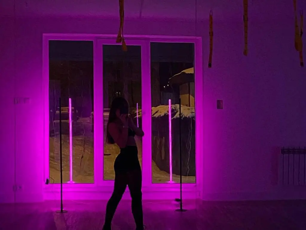 PaigePradoo poses in black lingerie in a room lit with bright pink neon lights near a large window.