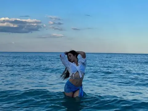 PaigePradoo — PaigePradoo posing in the ocean wearing a white shirt and blue bikini bottoms under a blue sky.