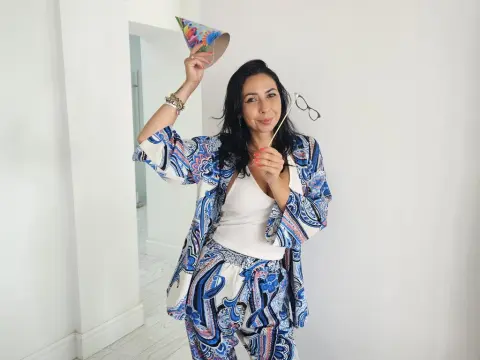 Cheerful woman in blue paisley loungewear holding a party hat and glasses in a bright white room.