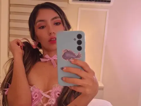 Young woman in pink lace lingerie and hair bows taking a selfie in a cozy bedroom setting.