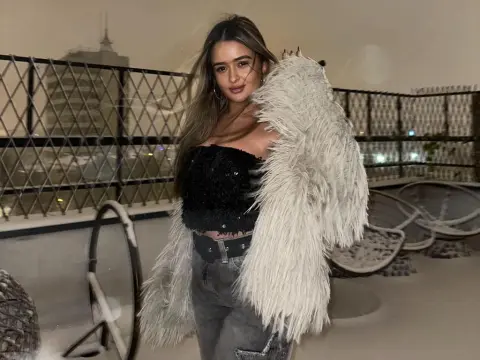 Young woman in a fuzzy white coat and black crop top posing on a rooftop terrace at night.