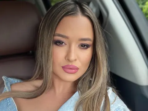 Young woman with long brown hair and glamorous makeup wearing a denim shirt sitting in a car.