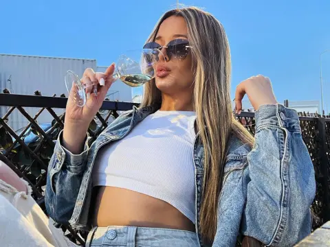 Blonde woman in denim jacket and white crop top sipping wine on a sunny rooftop terrace.