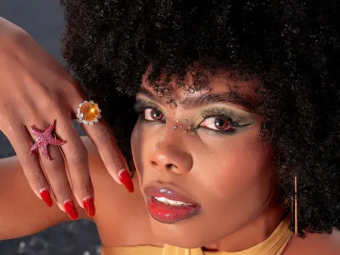 Woman with curly hair and glamorous makeup wearing statement star and gemstone rings in a moody studio setting.