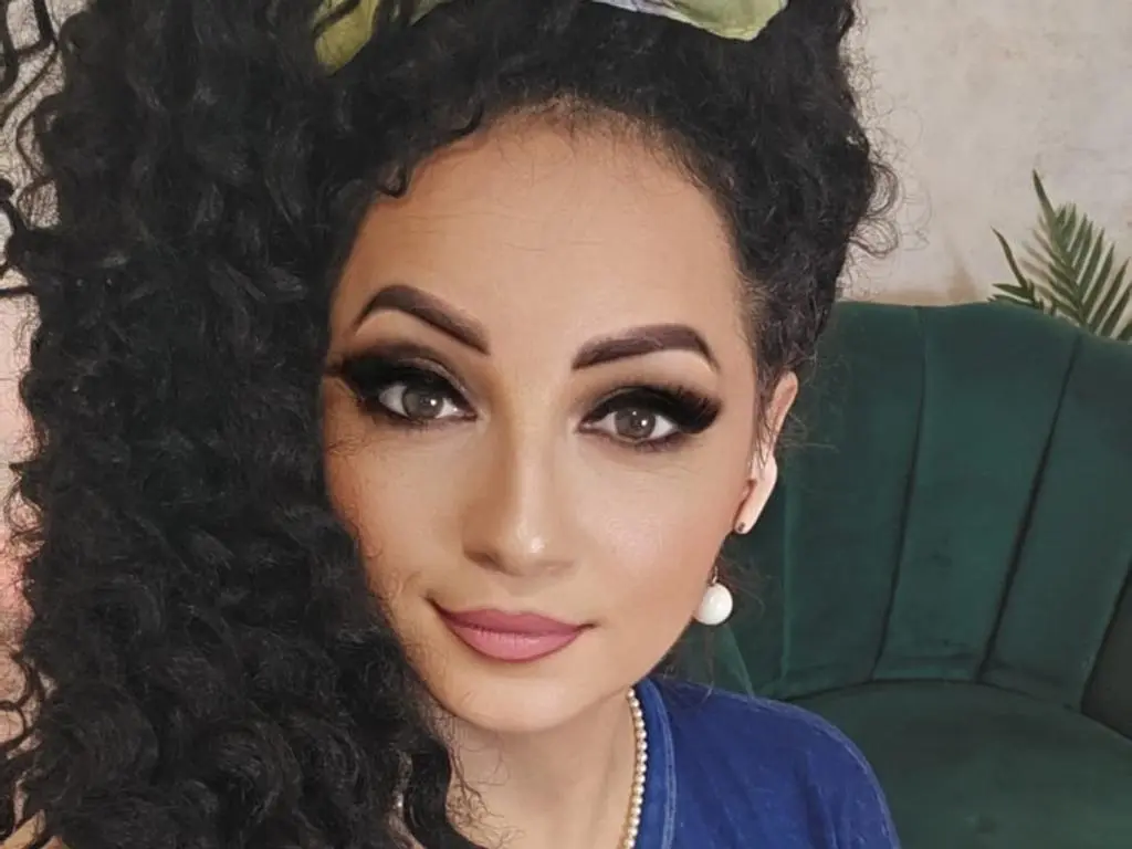 AnissiaTeasser — Woman with voluminous curly hair and glamorous makeup wearing a blue top and pearl earrings in a cozy setting.
